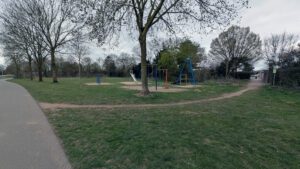 Ein kleiner Spielplatz auf einer Wiese, zwischen ein paar Bäumen. Links daneben der Fußgängerweg.