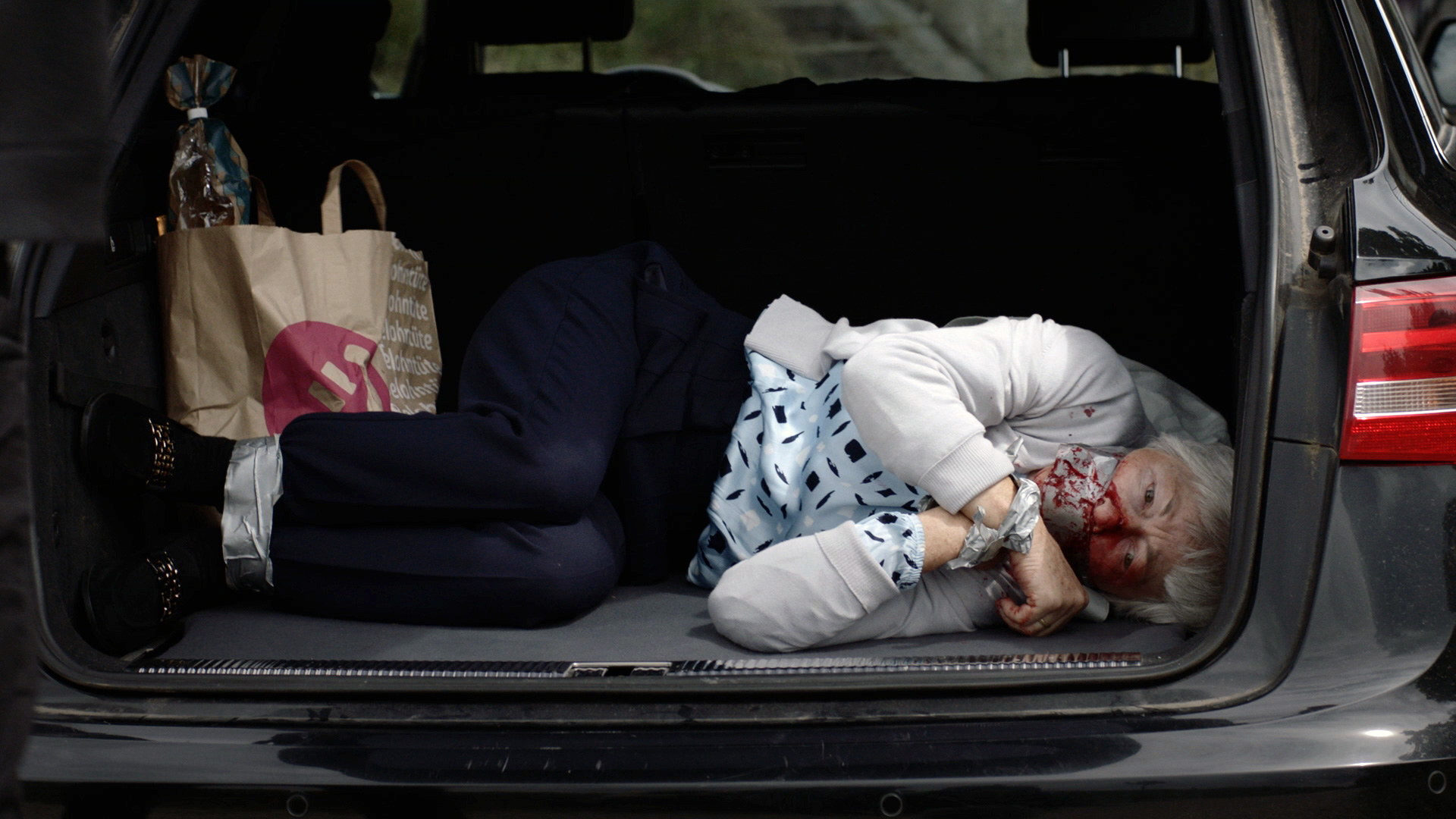 Eine Frau liegt im Kofferraum eines schwarzen Audis neben Einkaufstüten. Sie ist im Gesicht blutverschmiert und mit Panzertape gefesselt und geknebelt.
