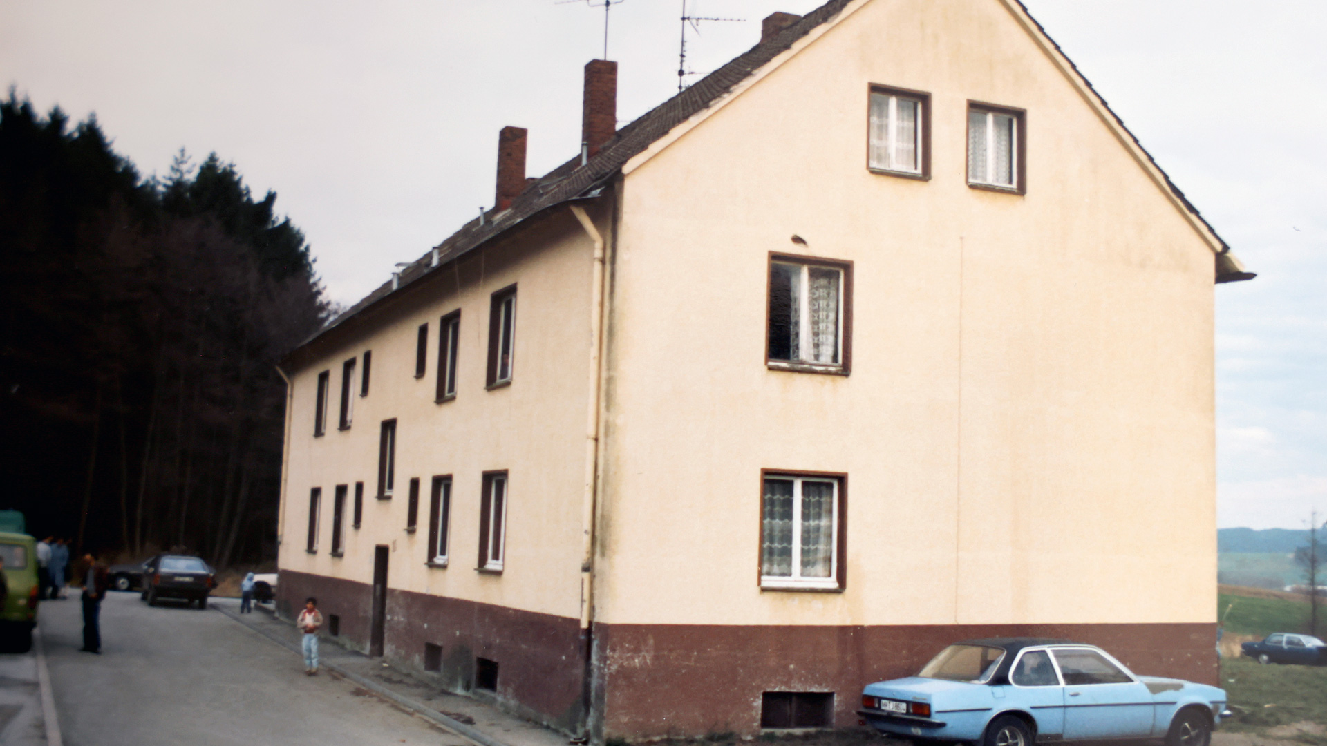 Ein Mehrparteienhaus aus den 80er Jahren, gelb gestrichen mit braunem Sockel und Satteldach.