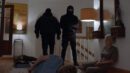 Im Flur eines Wohnhauses im Obergeschoss stehen zwei schwarz maskierte Männer, einer davon mit Pistole. Der Familienvater liegt mit Kabelbindern gefesselt am Boden, neben ihm kauert seine Frau, ebenfalls gefesselt.