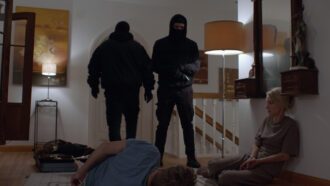 Im Flur eines Wohnhauses im Obergeschoss stehen zwei schwarz maskierte Männer, einer davon mit Pistole. Der Familienvater liegt mit Kabelbindern gefesselt am Boden, neben ihm kauert seine Frau, ebenfalls gefesselt.