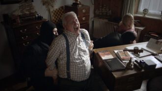 Zwei maskierte Männer fesseln einen Mann mit grauen Haaren und eine Frau an Stühle. Der Gefesselte schreit vor Schmerz auf.