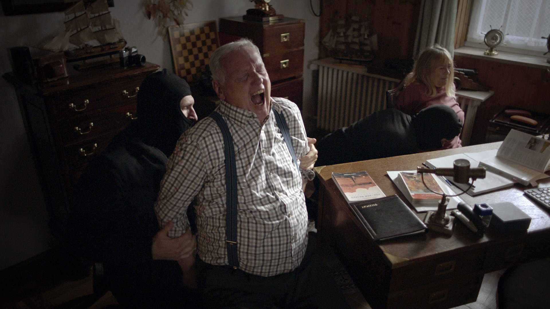 Zwei maskierte Männer fesseln einen Mann mit grauen Haaren und eine Frau an Stühle. Der Gefesselte schreit vor Schmerz auf.