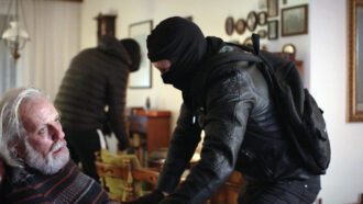 Ein schwarz gekleideter Räuber bedroht einen alten Mann. Im Hintergrund durchsucht ein weiterer Täter einen Schrank.