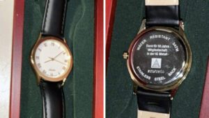 Vorder- und Rückseite einer Armbanduhr mit schwarzem Band, goldener Lünette und weißem Ziffernblatt in einer roten Box mit grünem Futter.