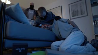 Ein Mann liegt blutüberströmt auf einer Couch. Hinter ihm stehen zwei weitere Männer, einer davon fasst ihn grob an der Schulter. Auf dem Boden liegen viele Gegenstände verstreut, darunter eine Schachtel mit der Aufschrift „Tag Heuer“.