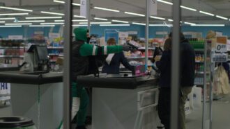 Durch die Fensterscheibe eines Discounters ist ein Mann in grünem Jogginganzug und schwarzer Weste zu sehen. Mit gezogener Waffe bedroht er die Angestellten.