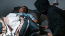 Eine ältere Frau liegt schlafend in ihrem Bett. Ein Mann mit schwarzer Kapuze beugt sich über sie