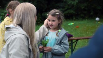 Ein kleines Mädchen mit Zöpfen steht auf einer Wiese. Sie hält einen Strauch grüner Blätter in der Hand. Eine erwachsene Person steht vor dem Mädchen kniet vor dem Mädchen und streichelt ihre Wange.