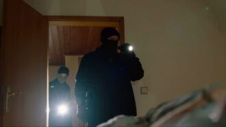 Zwei schwarz maskierte Männer mit Taschenlampen betreten ein dunkles Schlafzimmer.